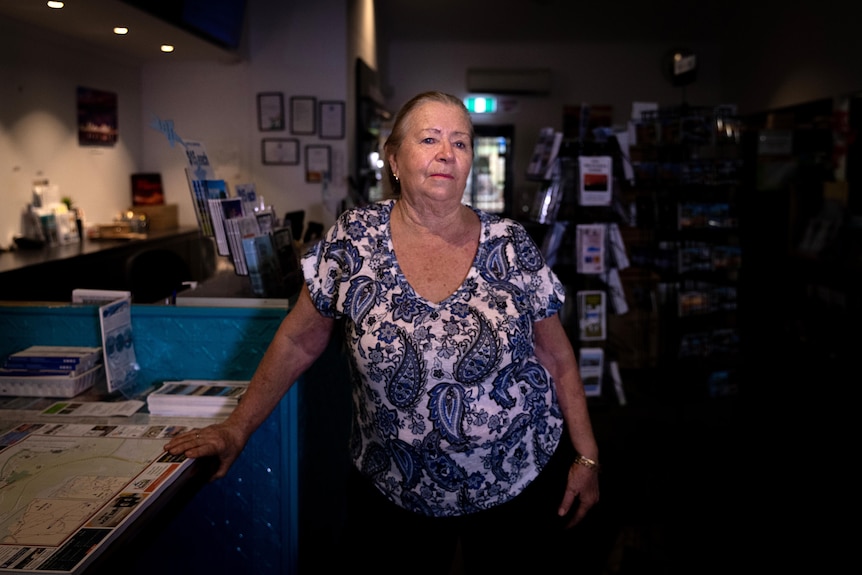 Ann older woman wearing a blue and white top stands inside a store with her hand on a counter and a serious look on her face.