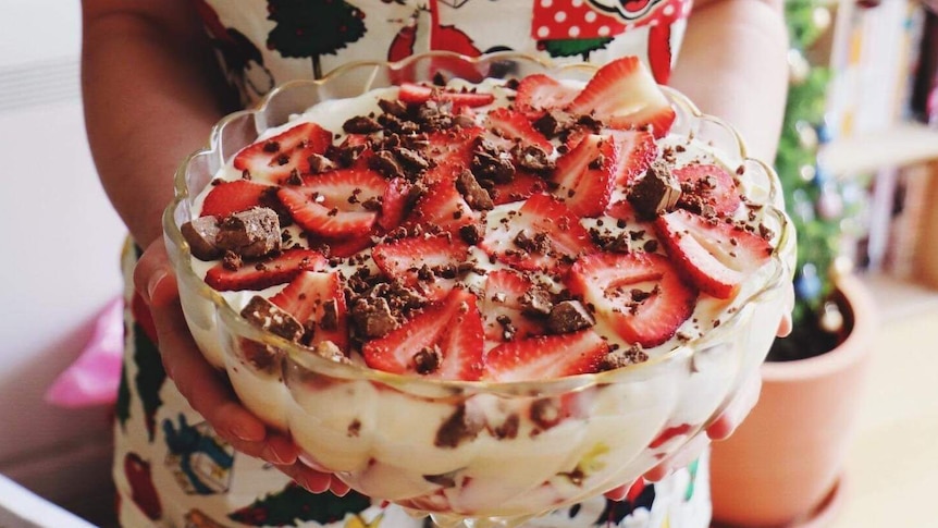 A woman holds a bowl of trifle topped with cream, chocolate and strawberries.