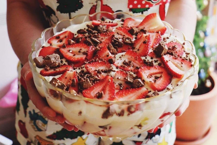 A woman holds a bowl of trifle topped with cream, chocolate and strawberries.