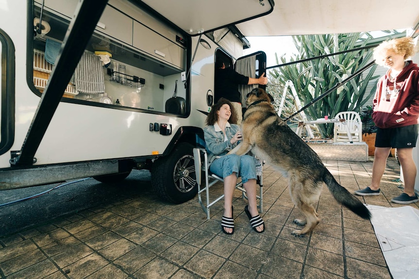 A woman and a dog play on a chair outside a caravan