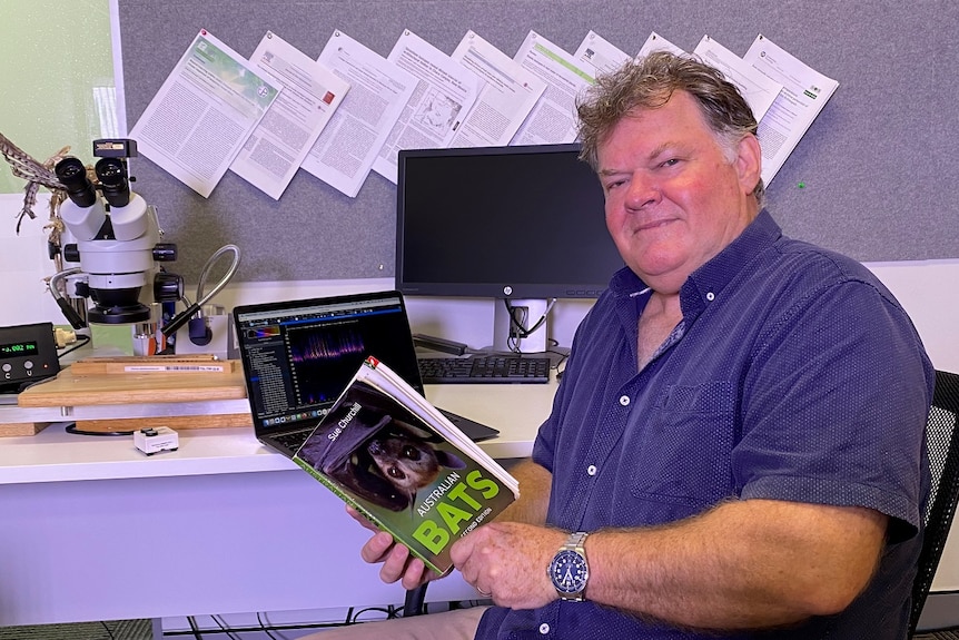 A middle-aged man sits at his desk and holds a book on bats.