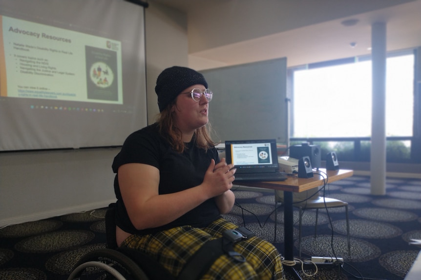 A woman in a wheelchair dressed in a black shirt with a beanie sits in a meeting room with a projecting screen behind her.