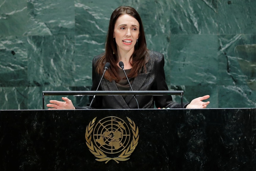 Jacinda Ardern stands behind a lectern with the UN logo on it, in front of a green marble wall