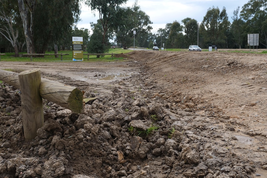 A dirt entrance to a campground blocked by a long dirt levee with a "Wilks Park" sign in the background next to a road.