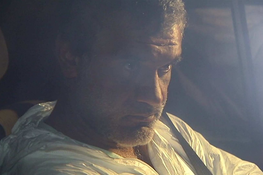 A man in a white single-use one-piece suit sits in the back of a car.