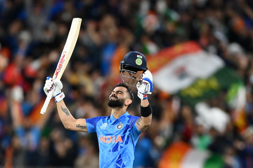 Virat Kohli holds his bat and helmet in the air and closes his eyes with India flags behind him
