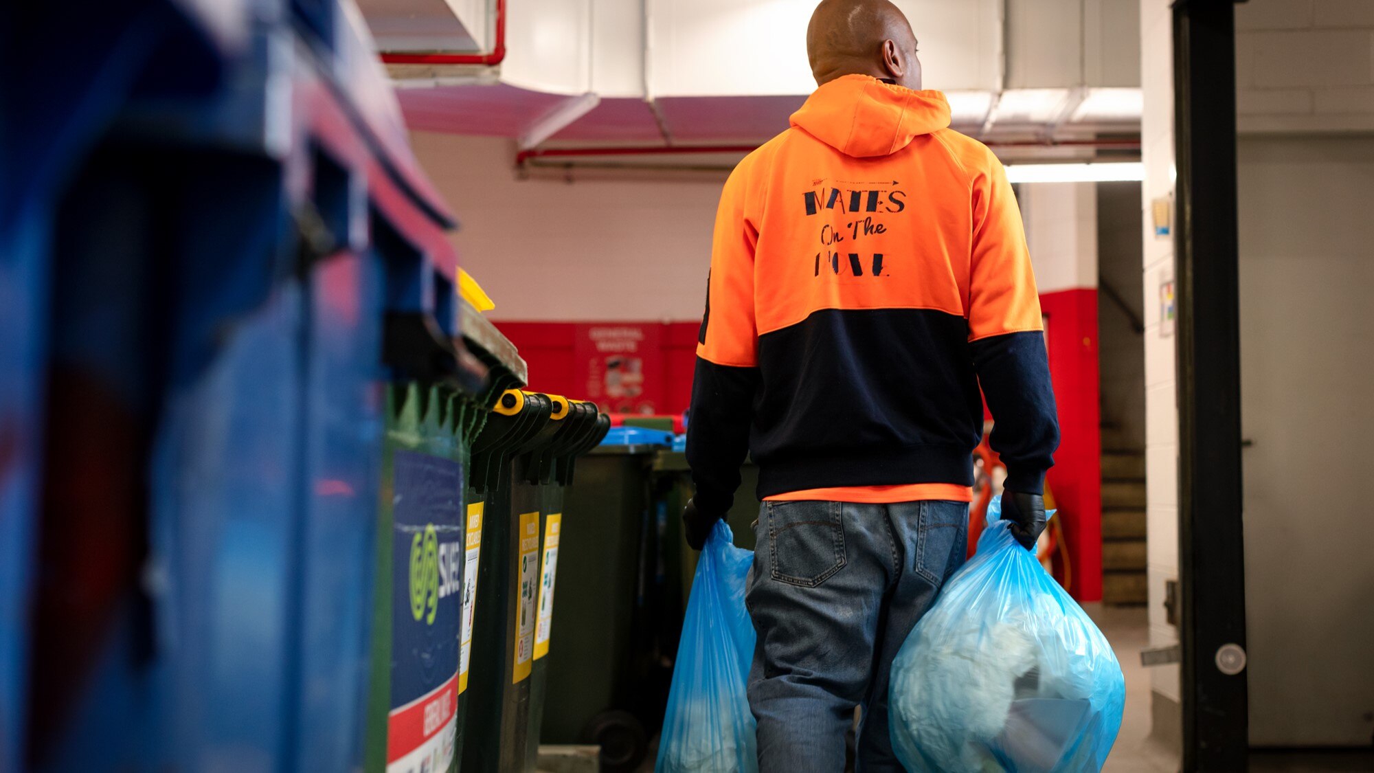 A man in a labeled orange hoodie "friends on the move" carries two trash bags next to some trash cans.