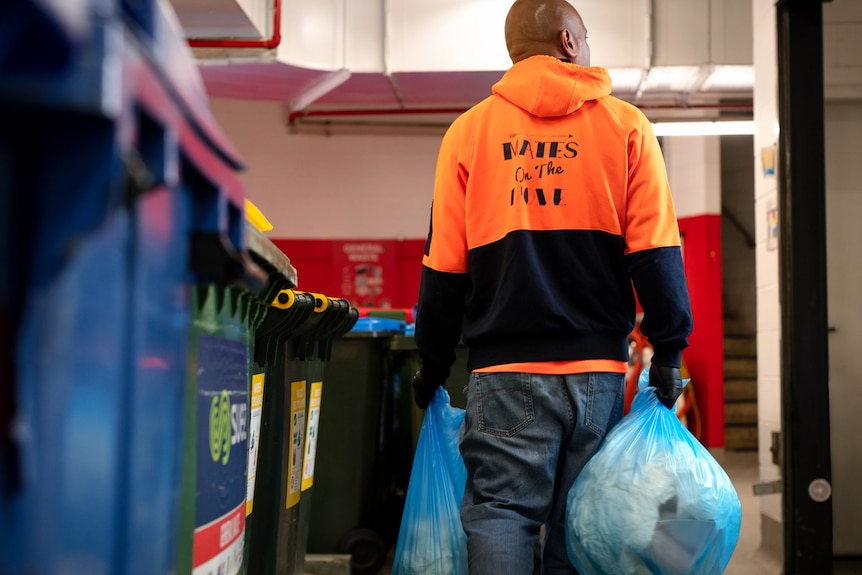 A man in an orange hoodie labelled "Mates on the Move" carries two rubbish bags alongside some bins.