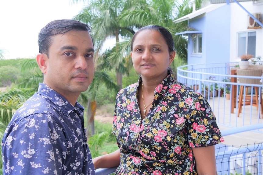 Damini and Santoush Patel look towards the camera, with a blue building and palm trees behind them.