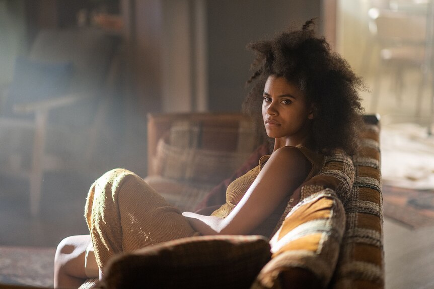 A young Black woman sitting on a 70s sofa in a loungeroom, sunlight pouring into the room