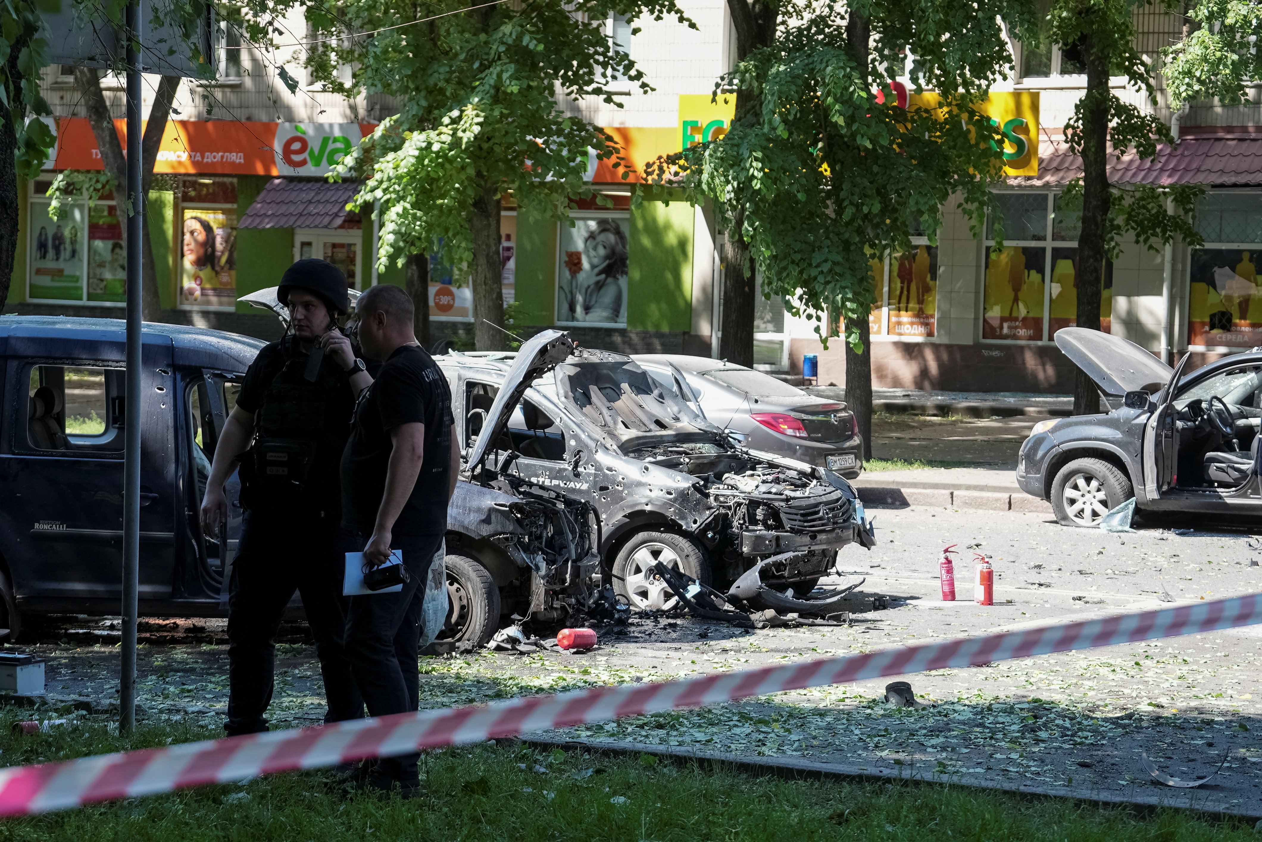 People stand near damaged cars behind police tape. 