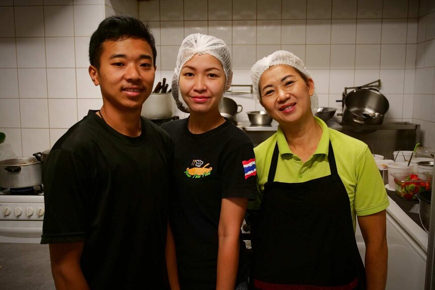 A man in a black shirt poses with two women wearing hair nets in a kitchen
