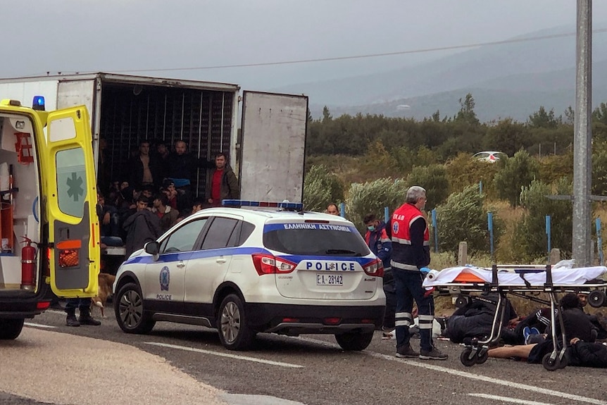 Migrants in the back of a truck, ambulance attending to people on the side of the road.