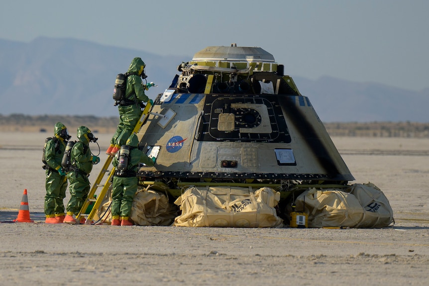 The NASA Starliner sits on the ground with cushions under it and men in hazard suits standing next to it