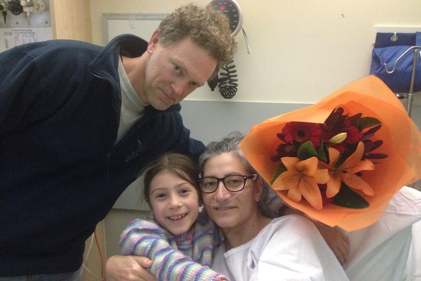 A woman in a hospital gown with a man and a young girl and a bunch of flowers.