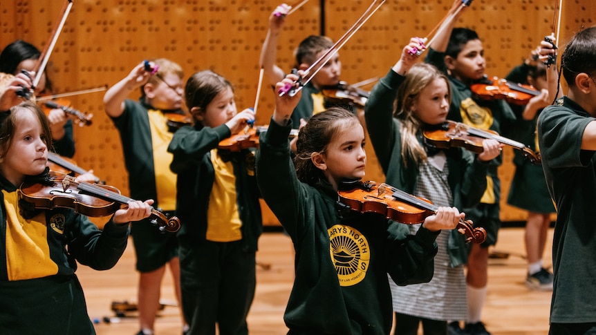 Primary school children in green and yellow uniforms play violins and hold their bows above their heads in a music auditorium.