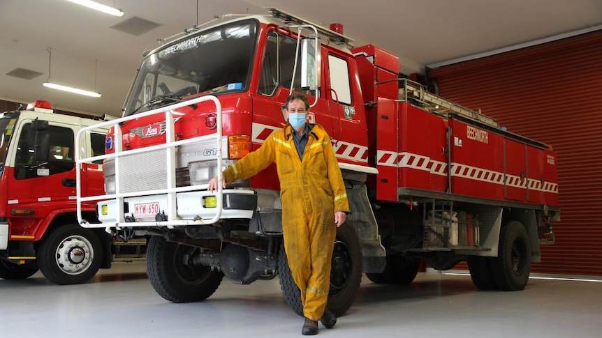 A man wearing yellow fire overalls and a face mask stands in front of a red fire truck.