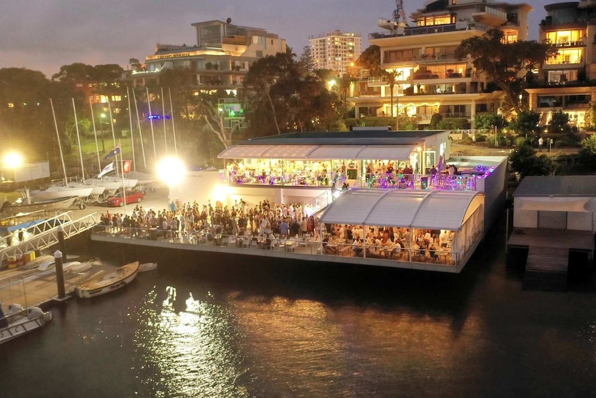 Wide shot of Cronulla Sailing Club at night with people in the club and on the deck overlooking the water