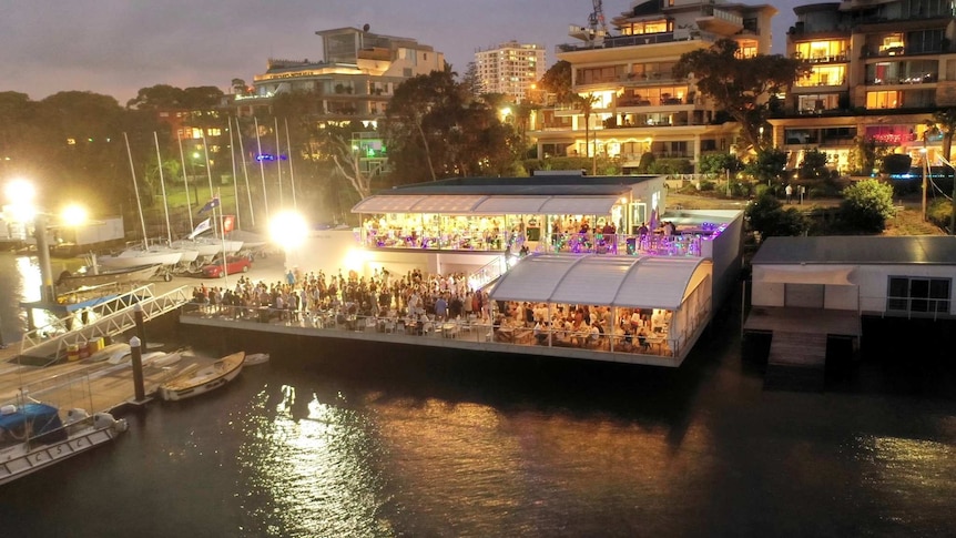 Wide shot of Cronulla Sailing Club at night with people in the club and on the deck overlooking the water