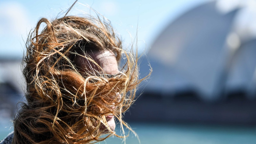 A woman's face is obscured by her hair being blown by the wind, Sydney Opera House is in the background