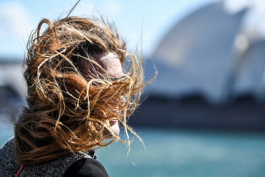 A woman's face is obscured by her hair being blown by the wind, Sydney Opera House is in the background