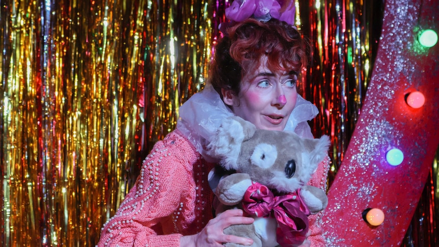 A woman in her late 30s in a pink clown costume holding a koala toy in front of a gold glitter backdrop