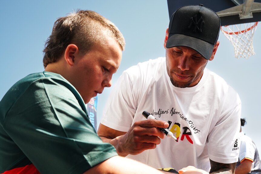 A young boy watching a man wearing a cap wear a pen about to sign merchandise.