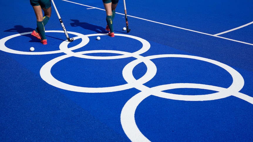 Two sets of legs standing with hockey sticks on a blue training field with the olympic rings in large white print.