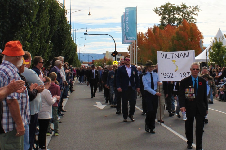 Veterans wearing suits and medals march down high street while a crowd look on. 