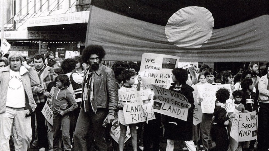 A black and white photo of an Aboriginal man with an afro in front of a protest with an Aboriginal flag.