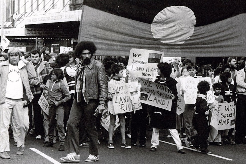 A black and white photo of an Aboriginal man with an afro in front of a protest with an Aboriginal flag.