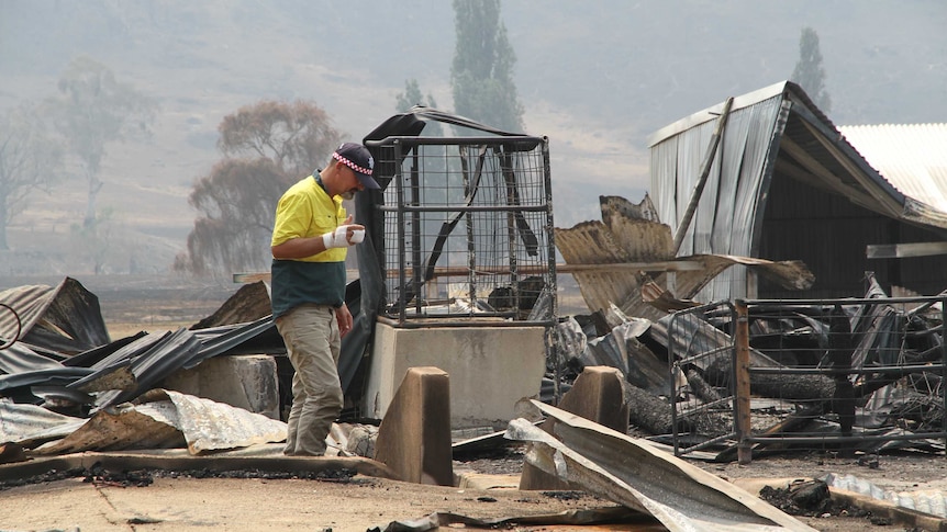 A man in a high-visibility yellow shirt and a bandaged hand walks through the rubble of a burnt house.