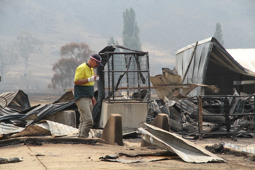 A man in a high-visibility yellow shirt and a bandaged hand walks through the rubble of a burnt house.