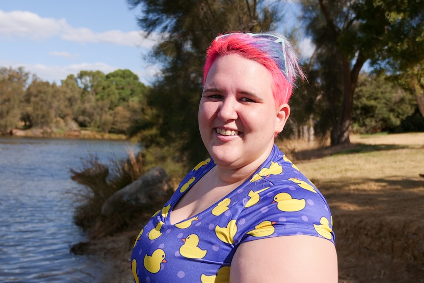 Woman with short pink and purple hair at riverbank with trees and water.
