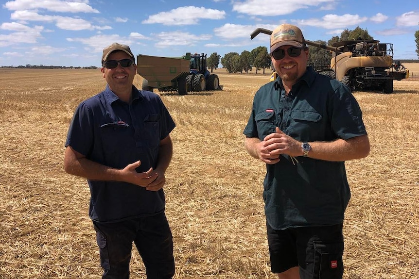 Two men stand in a paddock with a harvester and chaser bin behind them. 