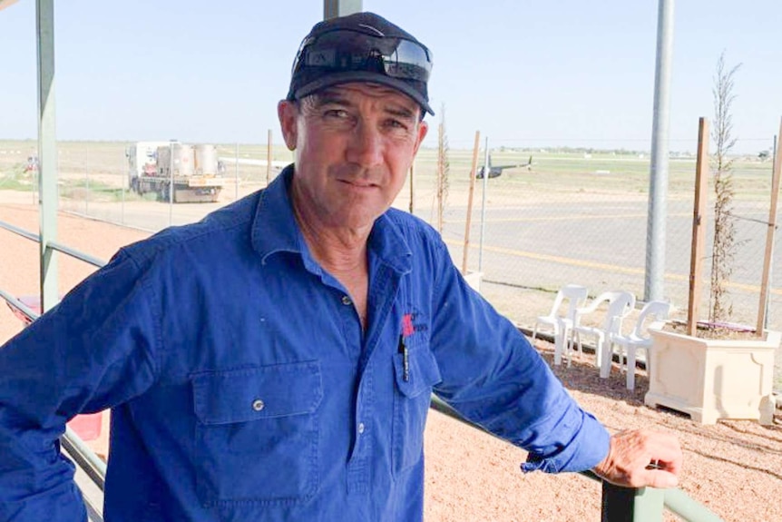 A man stands leaning on a deck railing, with a helicopter behind him.