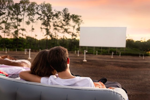 A man and a woman sit on a couch and are seen from behind as they look at a drive-in screen in the distance.