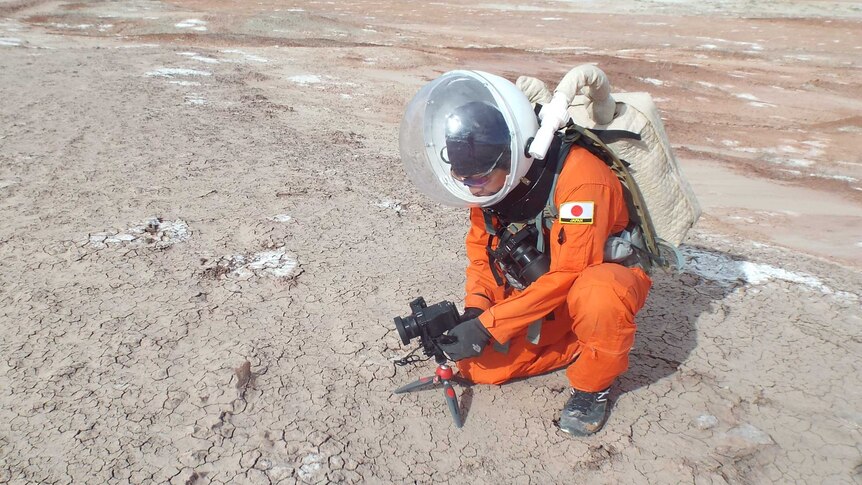 A man in an orange space suit sets up a camera in an arid landscape