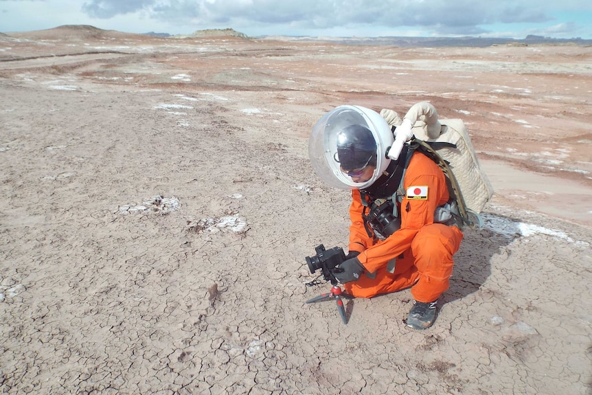 A man in an orange space suit sets up a camera in an arid landscape