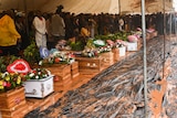 People stand behind a row of coffins surrounded by mud under a tent.