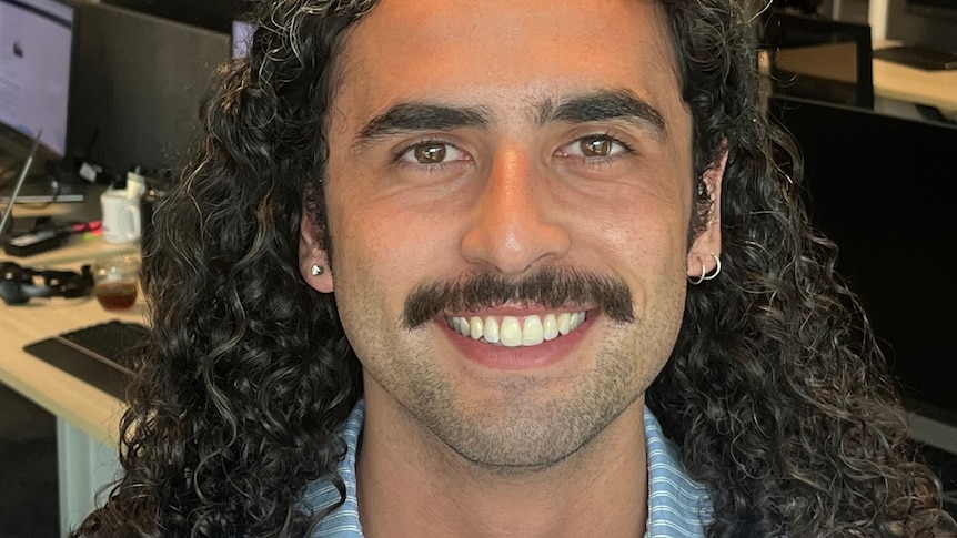 A man with long hair and a moustache smiles at the camera.