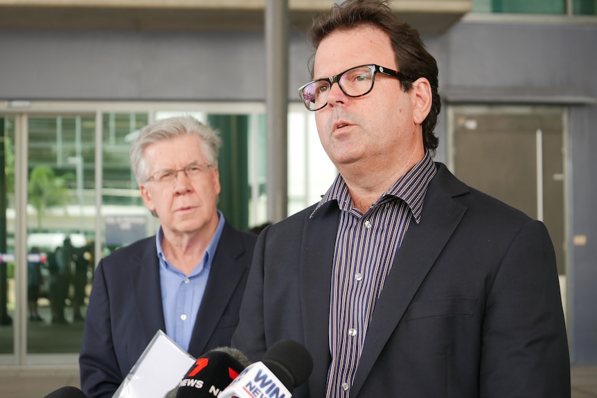 A man with dark hair and glasses speaks into a number of microphones. Behind him is an older man with white hair and glasses. 