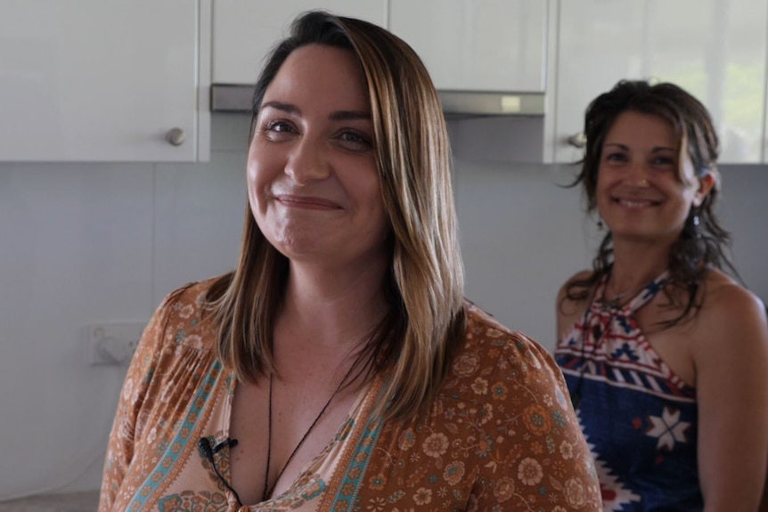A woman smiling to camera and a woman behind in a kitchen, also smiling, slightly out of focus.
