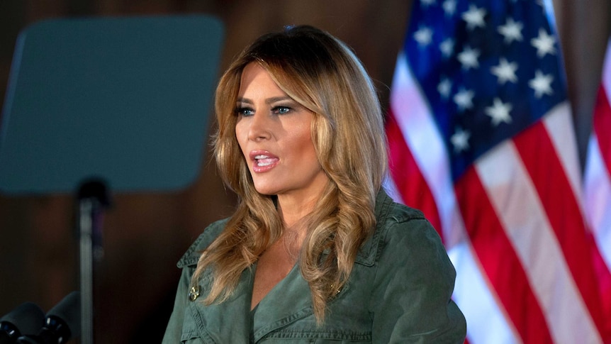 In a khaki-coloured dress, Melania stands in front of a microphone in front of US flags