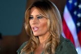 In a khaki-coloured dress, Melania stands in front of a microphone in front of US flags