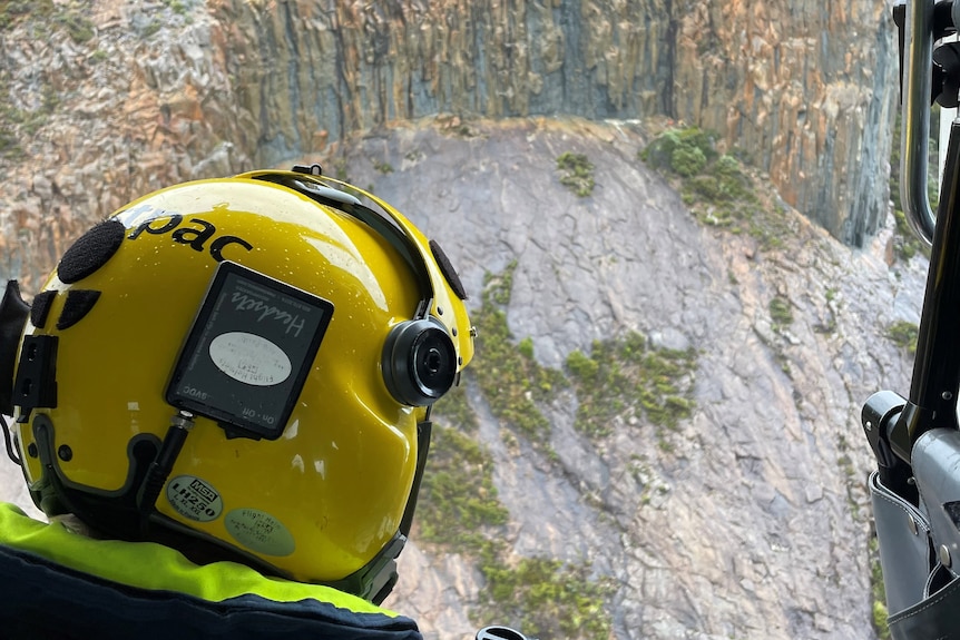 A view from a helicopter of a steep cliff face with the back of a police officer's helmeted head in the foreground