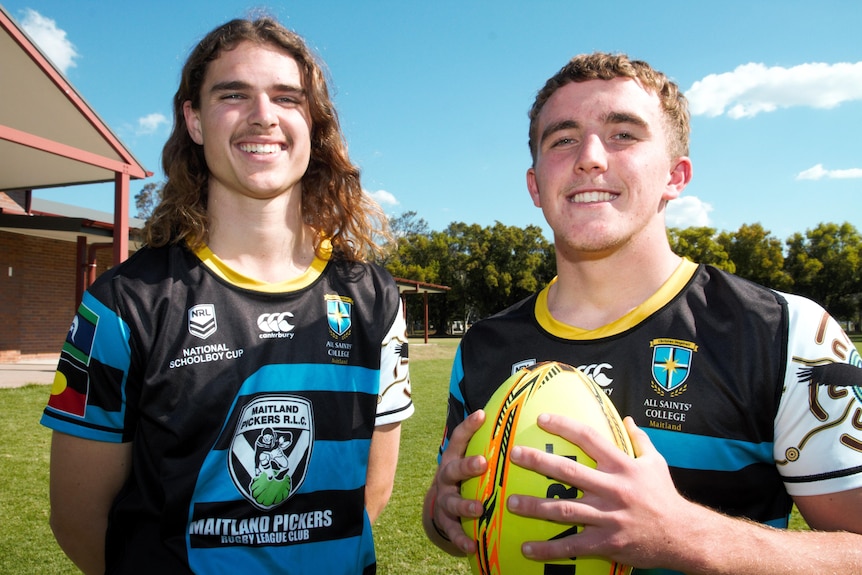 two teenage boys, students at a highschool, stand with a football. They're in their school's rugby league jersey