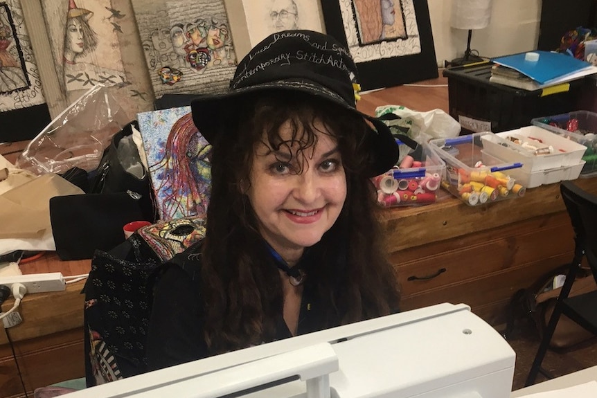 Dark-haired woman wearing hat smiling in front of white sewing machine