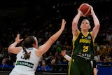 A tall blonde woman in a green and gold basketball jersey shoots a basketball over a tall brunette woman in a white jersey.