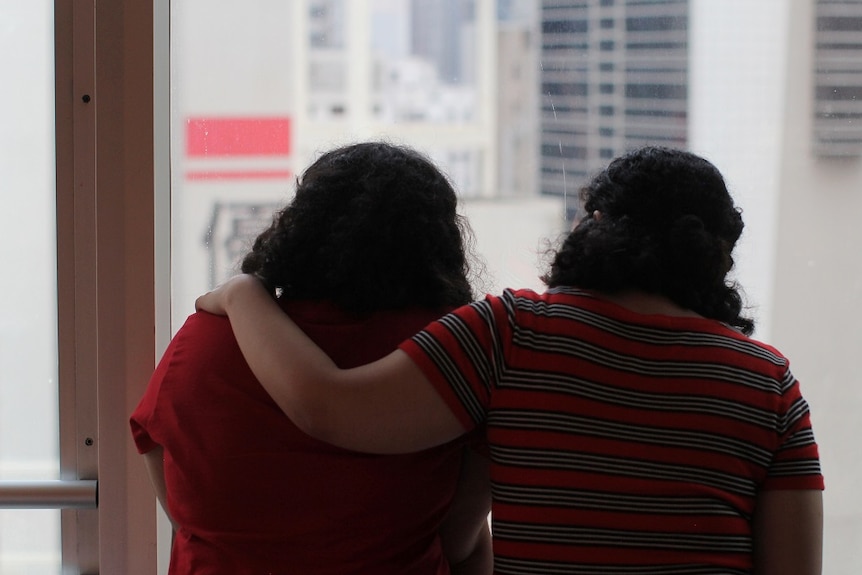 Two sisters, whose faces are not shown, sit together looking out a window while one places her arm around the other's shoulder.